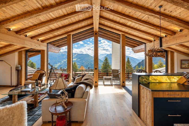 A spacious, well-lit living room with exposed wooden beams and large windows offering a scenic mountain view. The room features a cream-colored sofa, a glass coffee table, a wooden side table with a decorative deer head, and a modern kitchen island. Addit