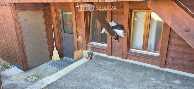 Wooden building exterior with a concrete pathway, broom, gloves, and a small potted plant near the entrance.