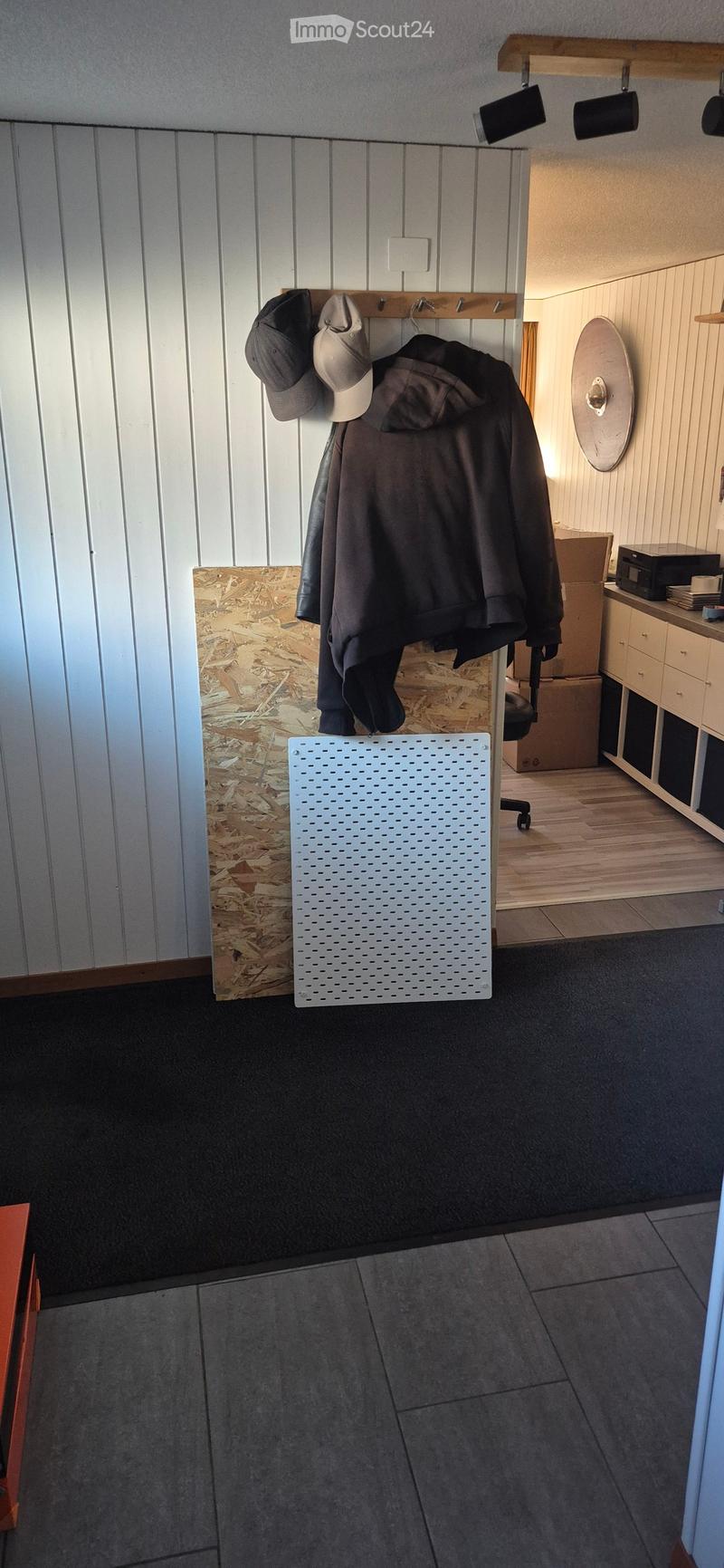 Hallway with white paneled walls, coat rack with jackets and hats, a white perforated board, and a small cabinet with drawers.