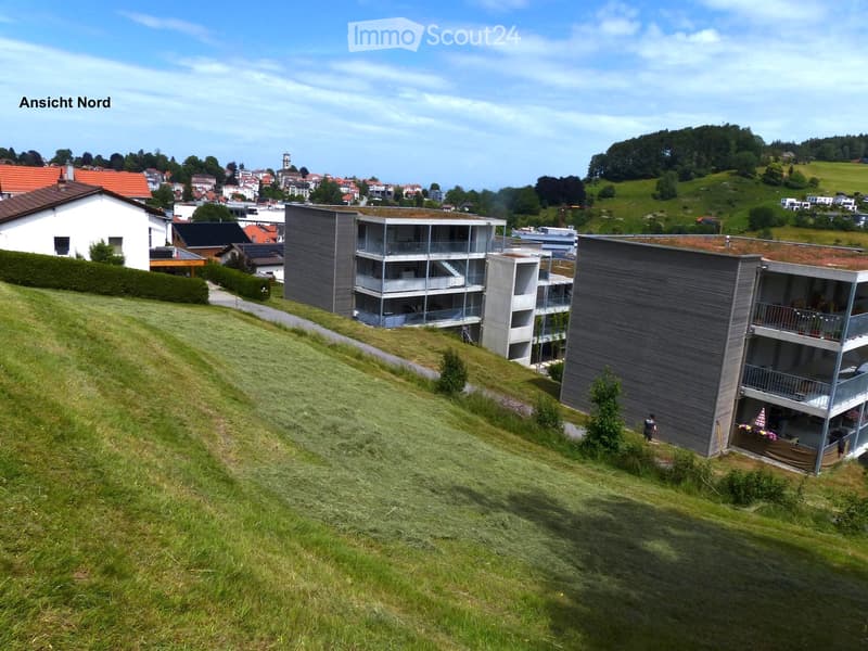 Modern multi-story residential building with balconies and green roof, situated on a grassy hillside overlooking a town with visible houses and a church tower.