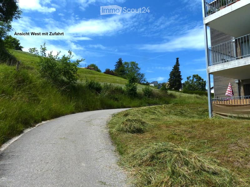 View of a residential building with a driveway, grassy slope, and a balcony with an umbrella visible on the right side.