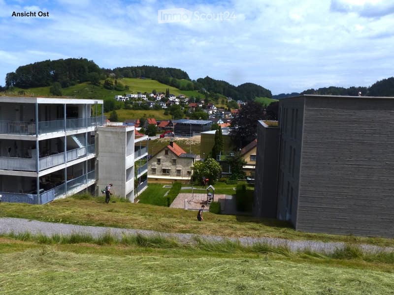 View of a modern residential complex with multiple buildings, including a multi-story apartment building with balconies, a smaller house with a red roof, and a contemporary structure with a dark facade. The area is surrounded by green hills and scattered