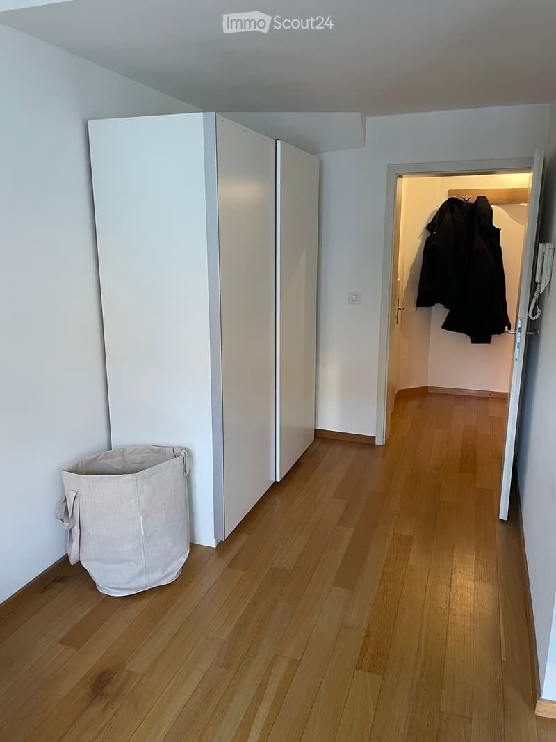 Empty hallway with light wooden flooring, white walls, and a partially open door leading to a closet area with coats hanging inside.