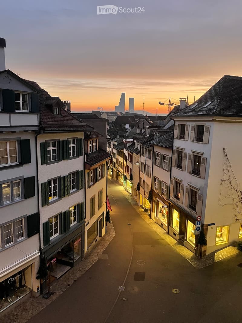 A narrow, cobblestone street lined with multi-story buildings featuring classic European architecture, illuminated storefronts, and a warm sunset sky.