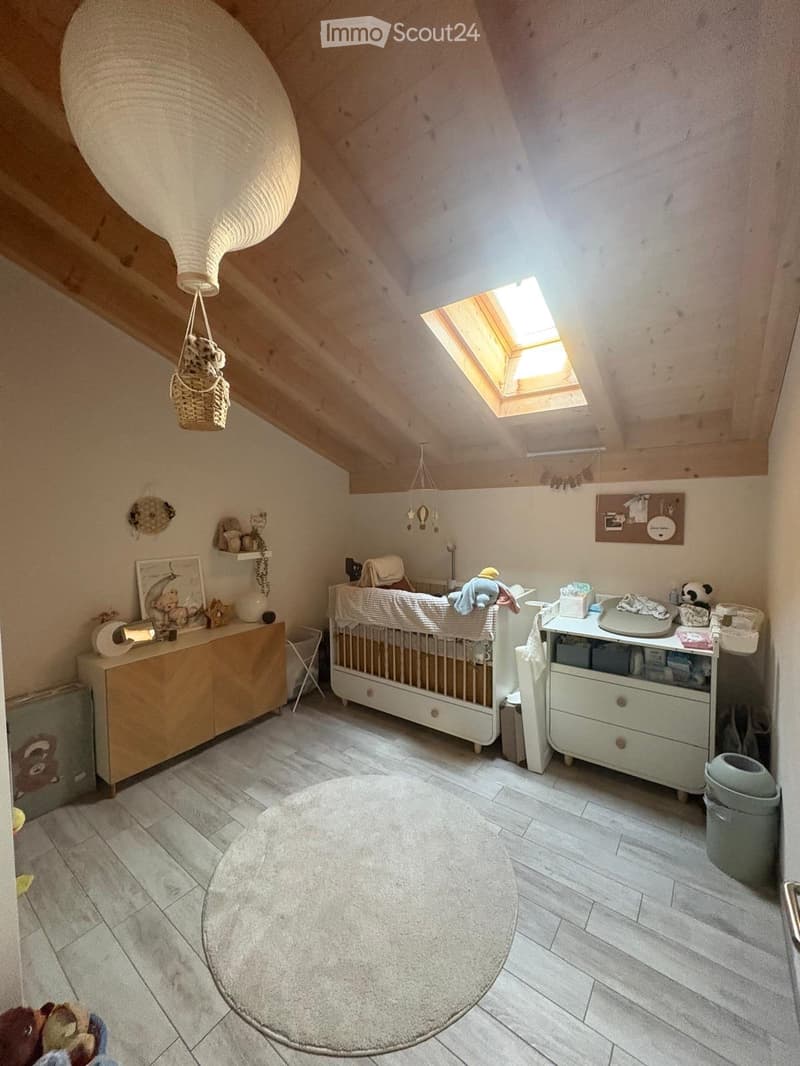 Cozy attic bedroom with wooden slanted ceilings, skylight window, crib, changing table, cabinet, and various baby items.