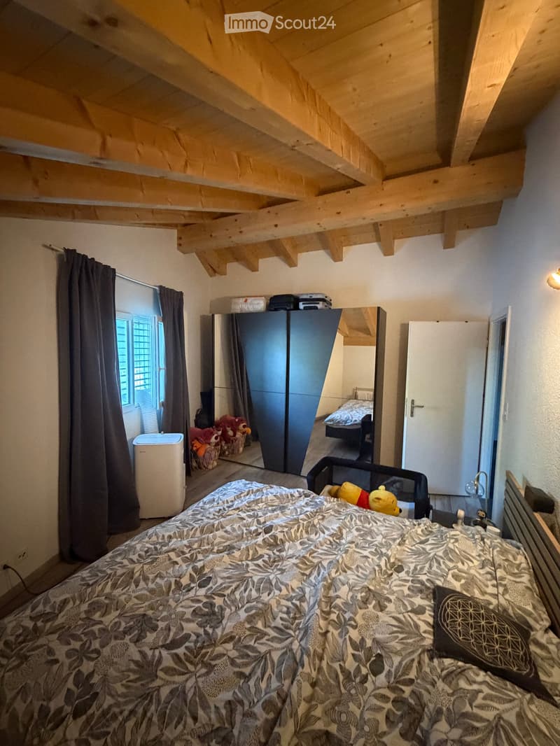 A cozy bedroom with a large bed, patterned duvet, and a mirror reflecting part of the room. The space features exposed wooden beams on the ceiling, a window with dark curtains, a wardrobe, and a small air conditioner unit near the window. Toys and a baske