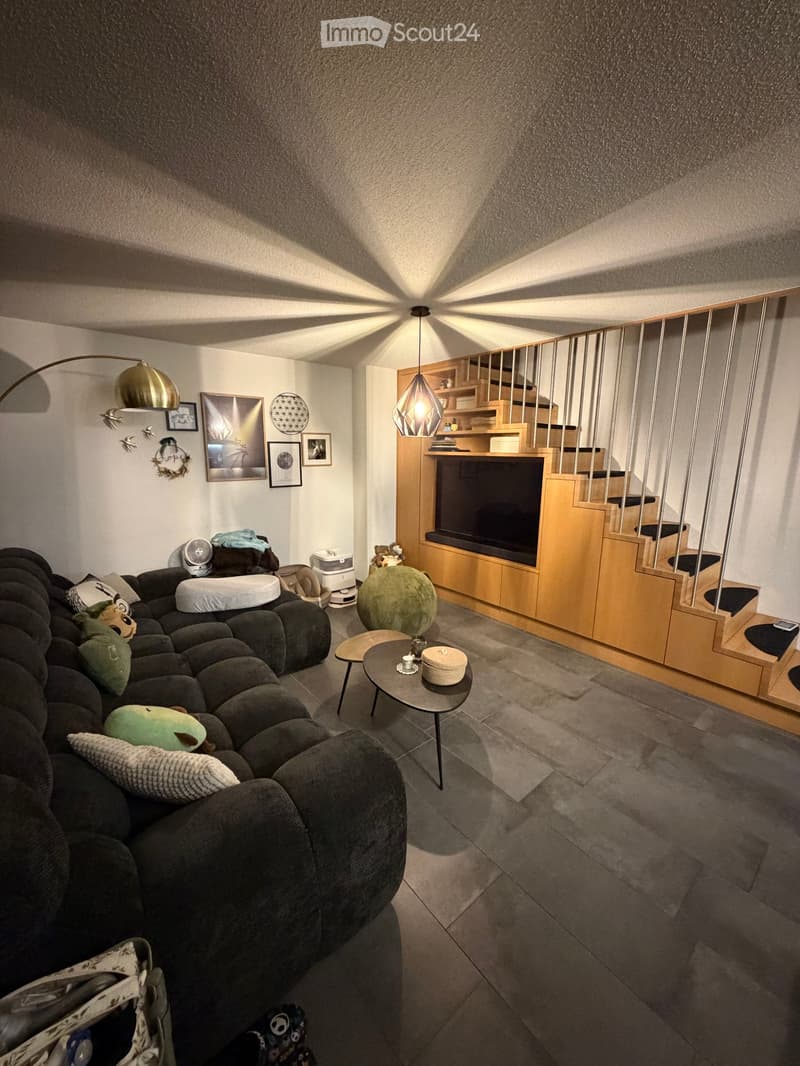 Modern living room with a dark grey sectional sofa, a round coffee table, a green ball chair, a staircase leading to an upper level, and decorative artwork on the walls.