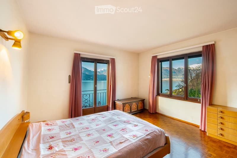 Bright bedroom with wooden flooring, large windows offering a scenic mountain and lake view, a bed with a floral quilt, a nightstand, and a chest of drawers.