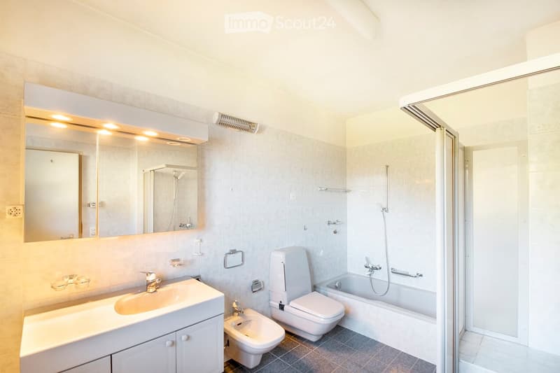 Modern bathroom with a large mirror, sink, toilet, bidet, bathtub, and a glass-enclosed shower. Light-colored tiles and warm lighting.