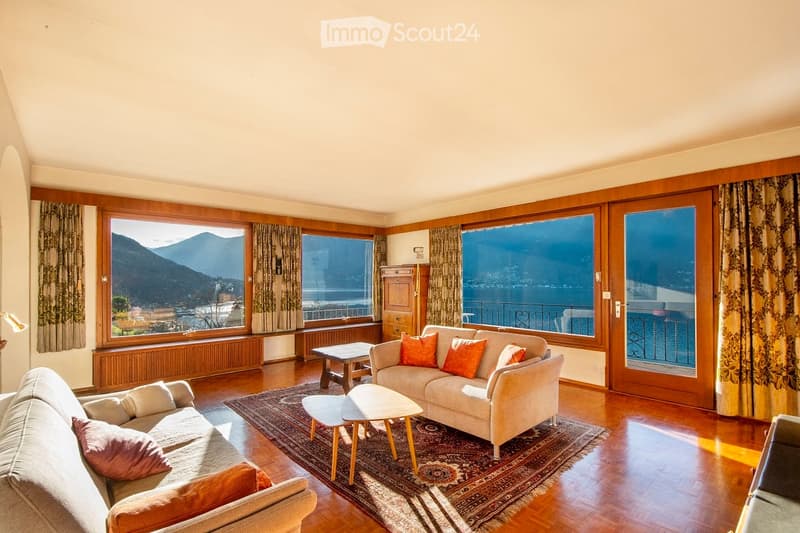 Bright living room with large windows offering a scenic mountain view, featuring a cream-colored sofa with orange cushions, a matching armchair, a coffee table, side tables, and a patterned rug on a wooden floor.