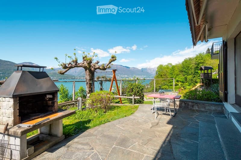 A serene outdoor garden area with a stone barbecue, a dining table with chairs, a swing, and a scenic view of a lake and mountains.
