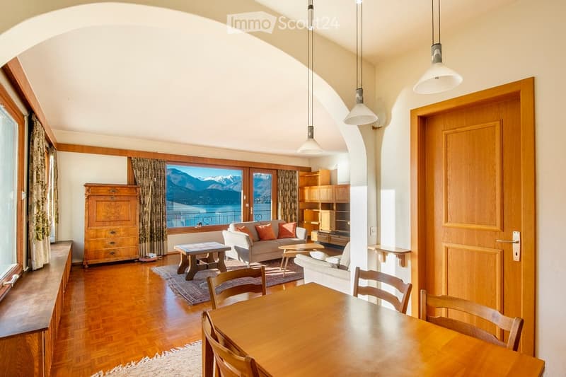 Bright living room with wooden floors, large windows offering a scenic mountain view, a sofa, coffee table, dining table, chairs, and built-in shelving.