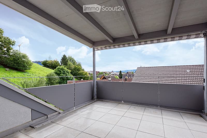 Modern balcony with tiled floor, grey walls, glass railing, retractable awning, and scenic view of greenery and rooftops.