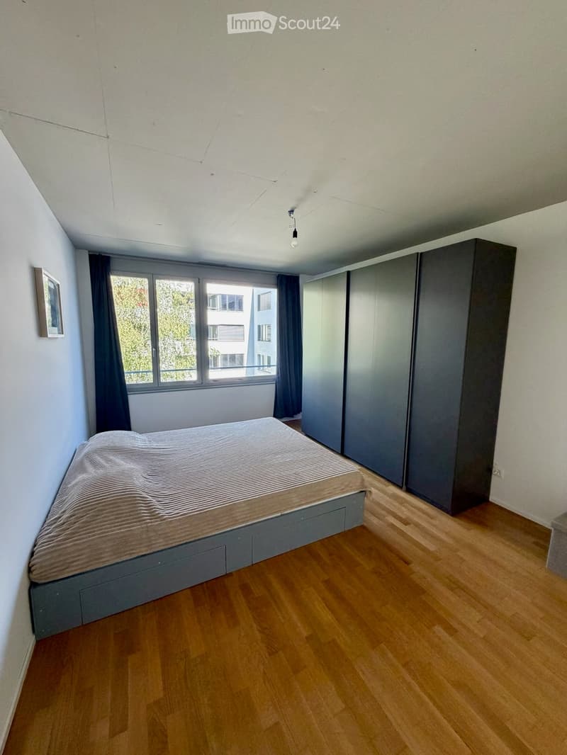 Modern bedroom with wooden flooring, a grey bed with striped bedding, a tall wardrobe, and large windows with dark curtains.