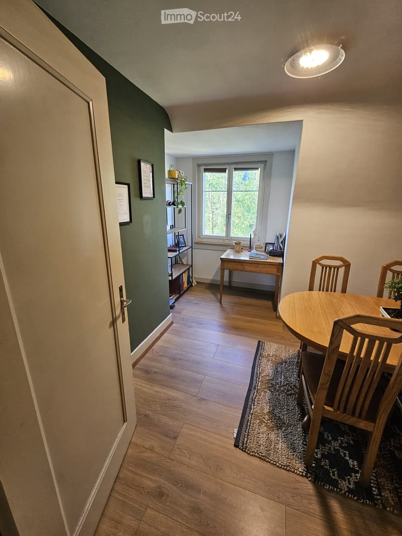 Dining area with wooden table, chairs, shelf, and desk, connected to a hallway with wooden flooring and a door.