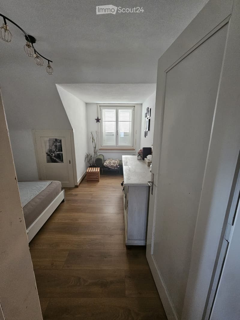 A furnished room with a bed, a small table, a chair, a counter, and a picture frame on the wall.