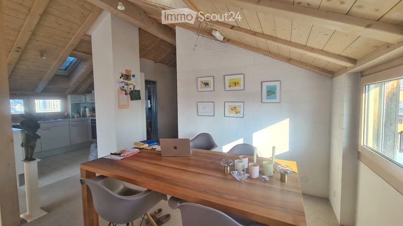 Bright, airy room with exposed wooden beams and white brick walls. Features a wooden dining table with chairs, a laptop, cups, candles, and framed pictures on the wall. A small kitchen area is visible in the background.