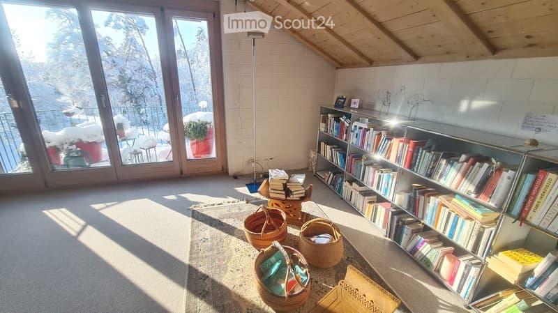 Bright, airy room with large windows offering a snowy outdoor view, featuring a bookshelf filled with books, several wicker baskets, a floor lamp, and a small table with chairs.