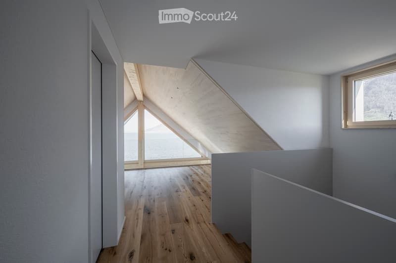 Bright, minimalist hallway with wooden flooring, white walls, and a skylight ceiling allowing natural light to flood the space. A window on the right side offers a view of the outdoors.