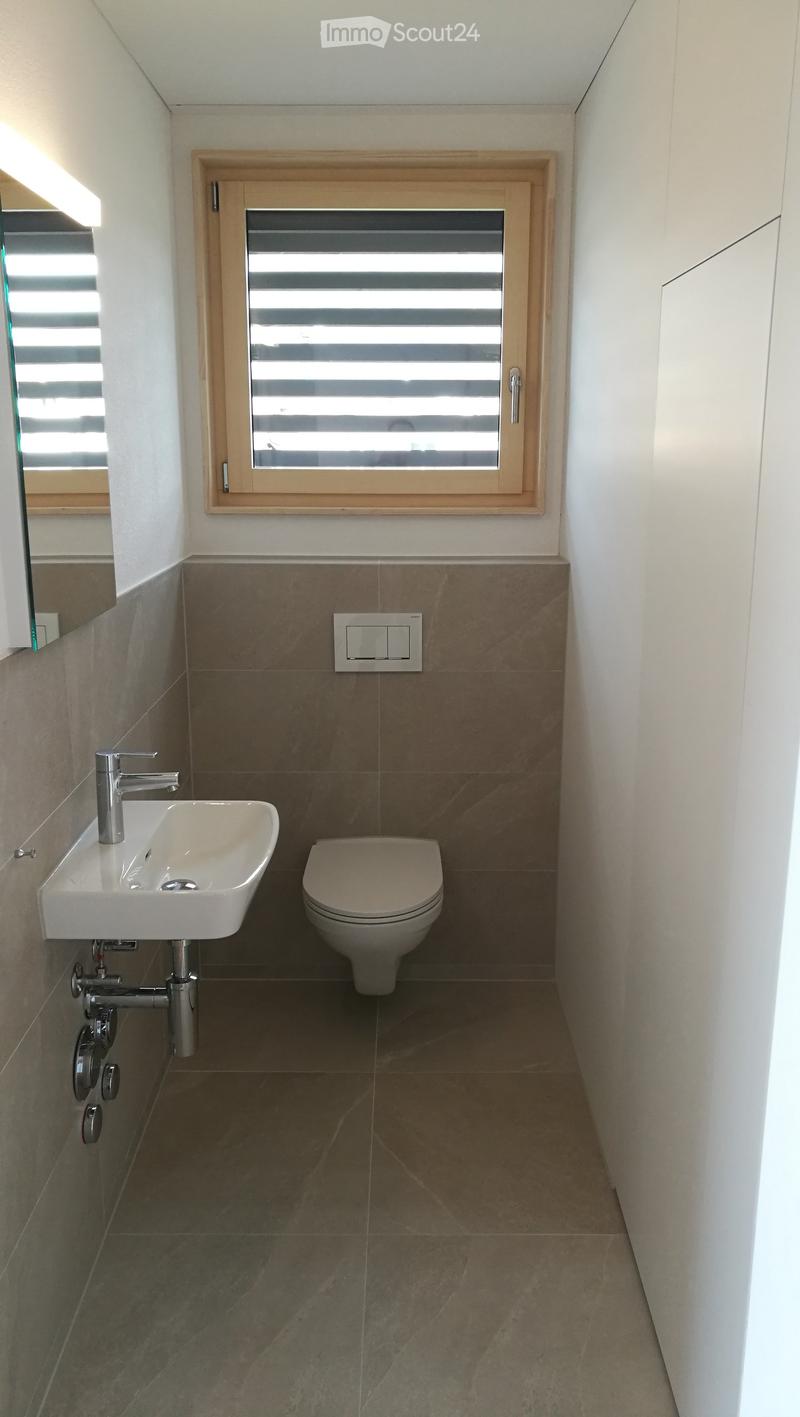 Modern bathroom with a white sink, chrome faucet, toilet, and a window with blinds. The walls are tiled in a two-tone design with brown lower tiles and white upper tiles.