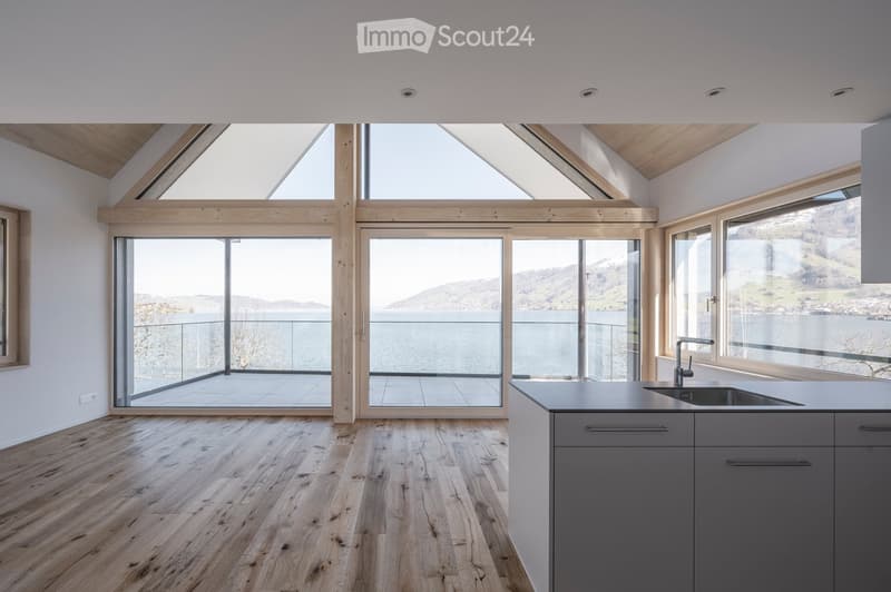 Modern kitchen with wooden flooring, grey cabinets, and large windows offering a scenic view of a lake and mountains.