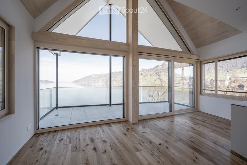 Bright, open loft-style room with wooden flooring, large glass sliding doors leading to a balcony, and expansive views of a lake and surrounding landscape.
