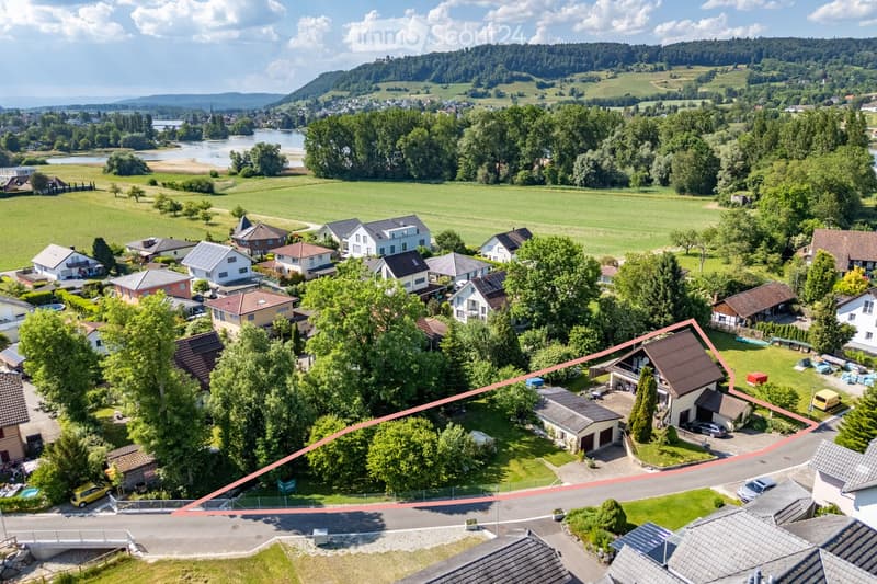 Aerial view of a residential neighborhood with multiple houses, surrounded by green fields, trees, and a river in the background.
