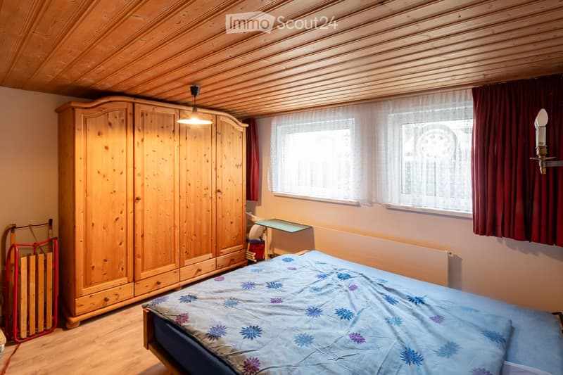 Cozy bedroom with wooden ceiling, large window with curtains, bed with blue floral duvet, wooden wardrobe, small table, and wall-mounted lamp.