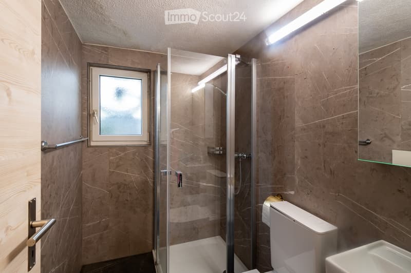 Modern bathroom with marble walls, a glass-enclosed shower, a toilet, a sink, and a mirror. Features include a frosted window and built-in lighting.