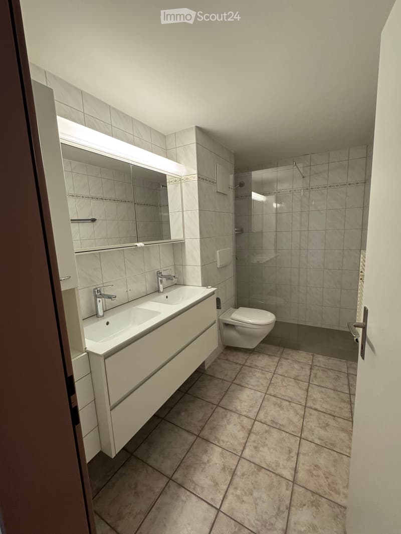 Modern bathroom with beige tiles, double sink, large mirror, and glass-enclosed shower area.