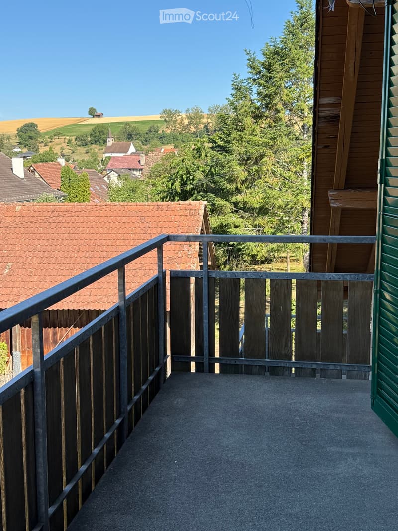 Balcony with wooden railing and metal balustrade, overlooking a rural landscape with houses, trees, and fields.