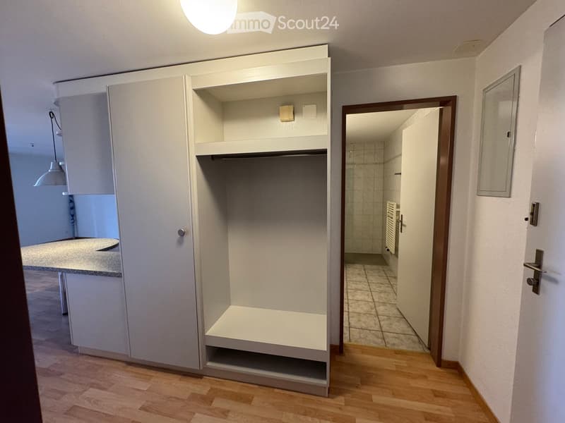 Empty hallway with wooden flooring, white walls, and a built-in closet with open shelving. A doorway leads to a tiled bathroom area.