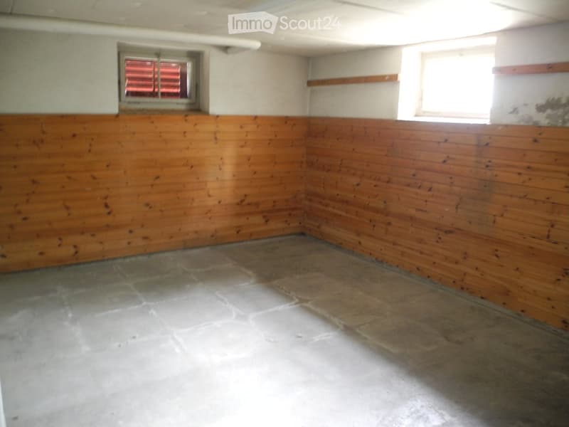Empty room with wooden panel walls and concrete floor, two windows with blinds, and exposed ceiling beams.