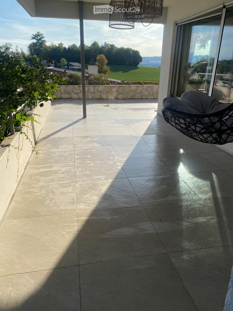 Balcony with tiled floor, wicker hanging chair, stone wall, and scenic countryside view.