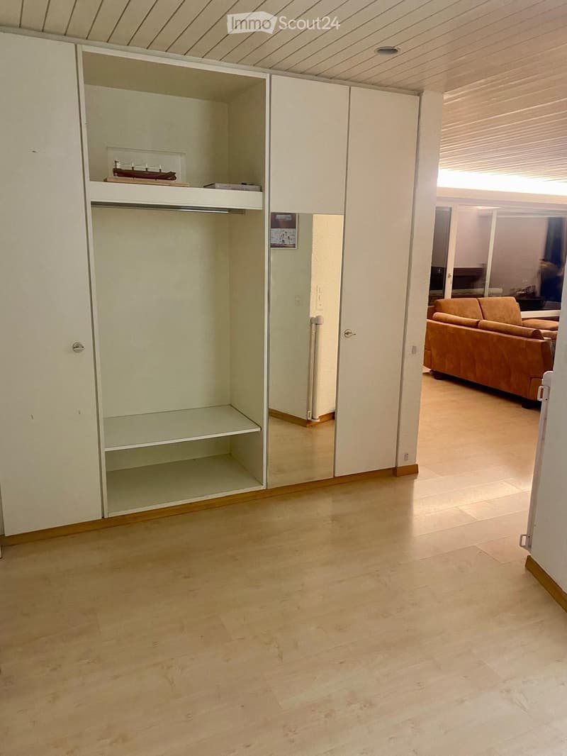 A furnished room with a wooden floor, a large closet with mirrored doors, and a visible living area with a sofa.