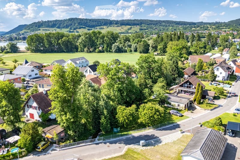 Aerial view of a residential area with multiple houses, surrounded by greenery and a scenic landscape with hills and a river in the background.