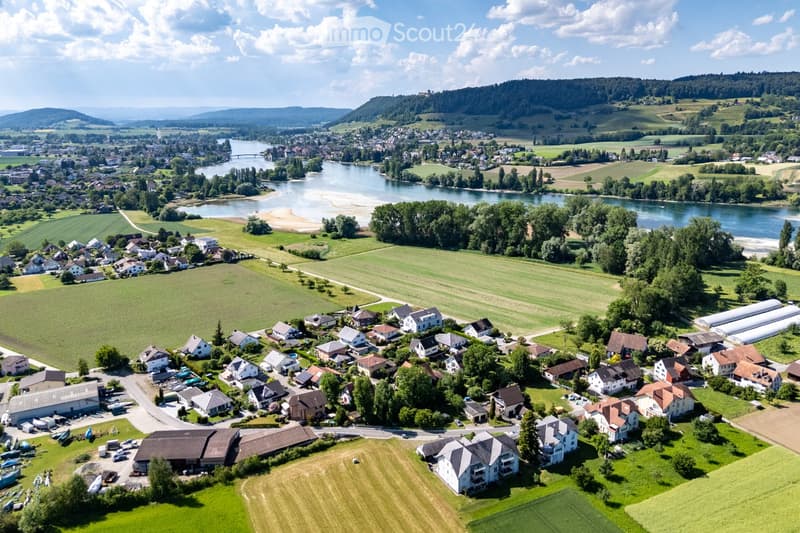 Aerial view of a small village with houses, fields, and a river running through it. The village is surrounded by green fields and trees, with a distant hill in the background.