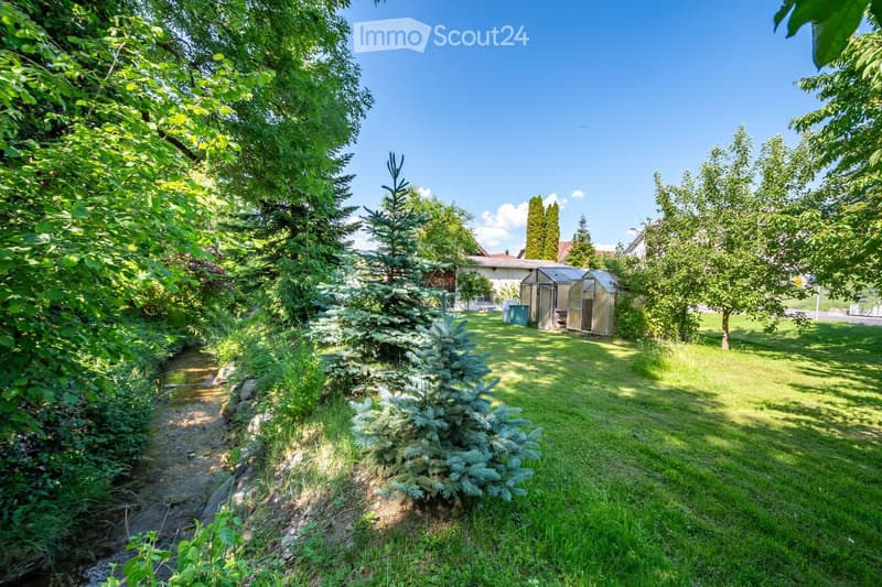 A well-maintained garden with a small stream, lush greenery, a greenhouse, and a house in the background.