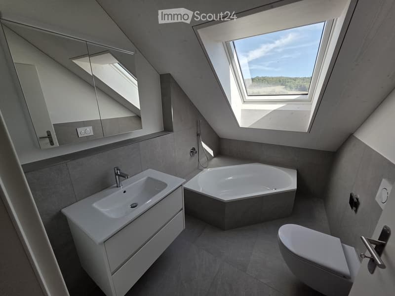 Modern bathroom with a sleek sink, bathtub, toilet, and mirrored cabinet, featuring large skylight windows and grey tiled walls.