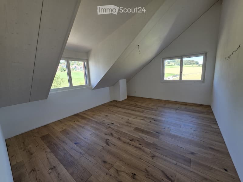 Empty attic room with wooden flooring, white walls, and large windows offering a view of the countryside.