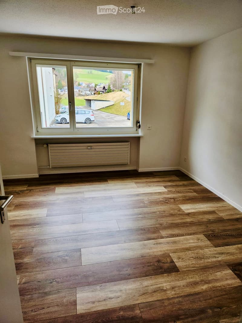 Empty room with wooden flooring, large window with view of the outside, radiator below the window, and neutral-colored walls.