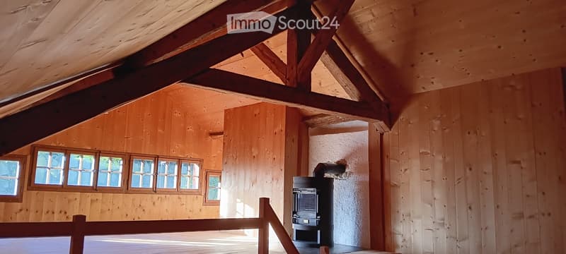 An unfurnished room with wooden walls and ceiling beams, large windows, and a black wood-burning stove.