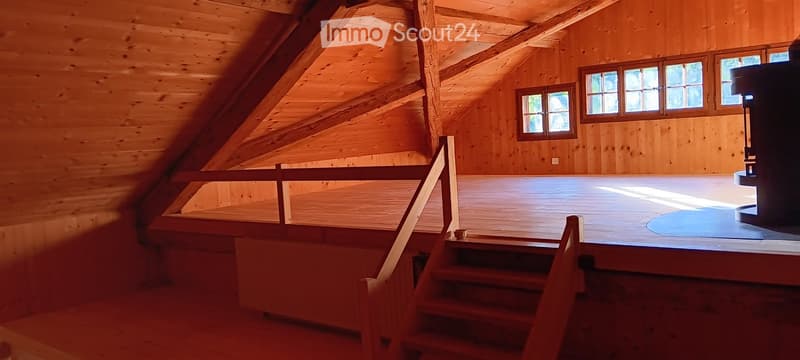 An empty attic space with wooden walls, exposed beams, and a staircase leading to a landing area. The room has large windows allowing natural light to enter.