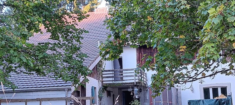 Traditional two-story building with a tiled roof, white walls, and a small balcony on the upper floor, surrounded by trees.