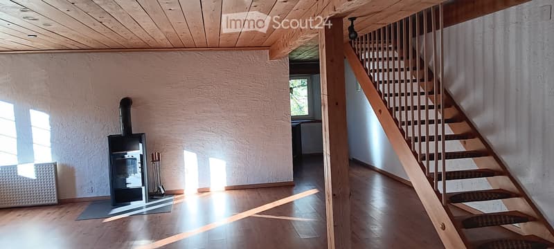 Modern hallway with wooden flooring, exposed wooden beams on the ceiling, a staircase leading upwards, and a black wood-burning stove with a chimney on the left side.