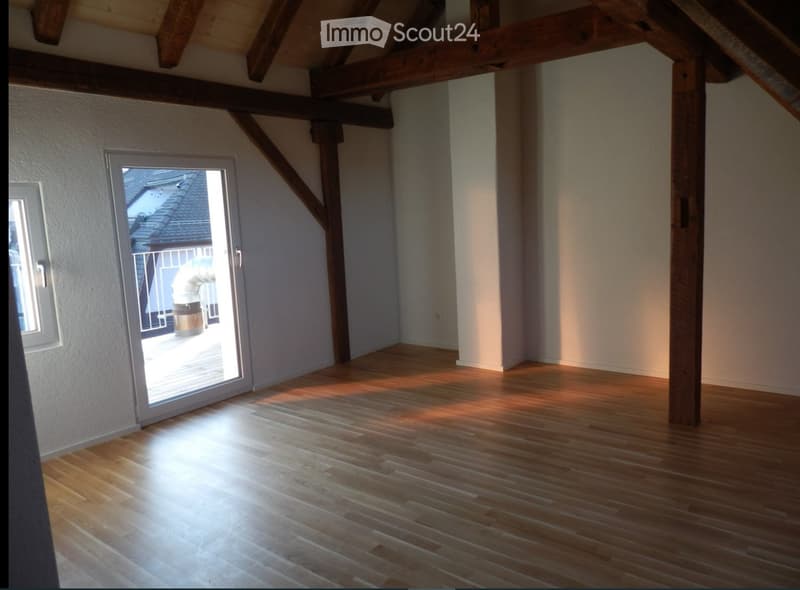 Empty room with wooden flooring, exposed wooden beams, and a sliding glass door leading to a balcony.