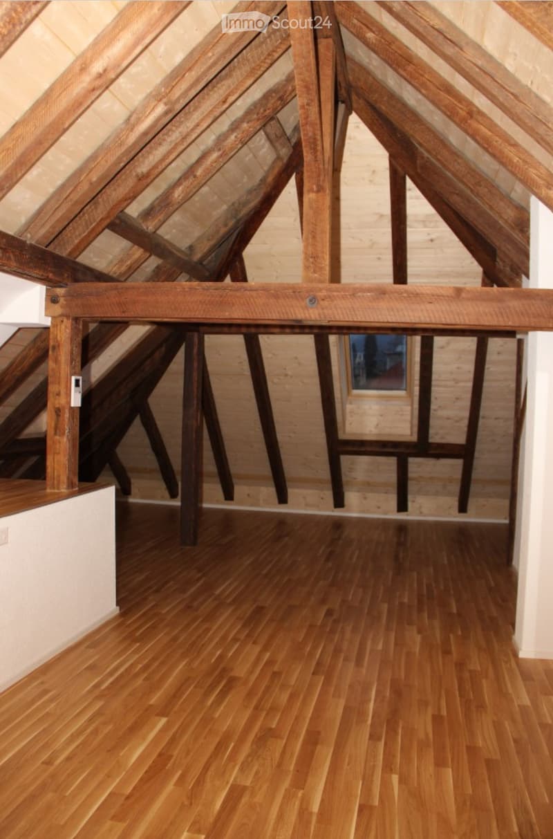 An attic space with exposed wooden beams and a polished wooden floor, featuring a staircase leading up and a small window at the far end.