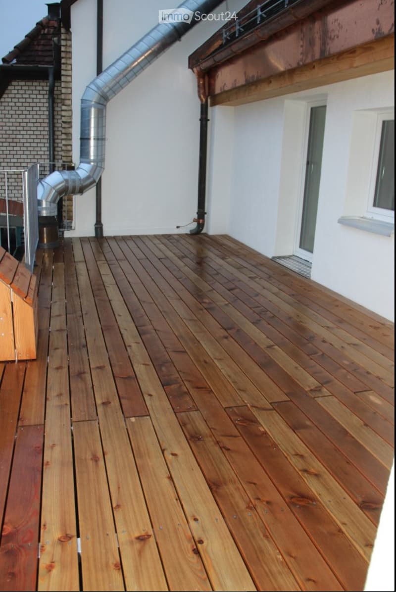 Wooden deck with a metal railing, white walls, and a metal pipe running along the side.
