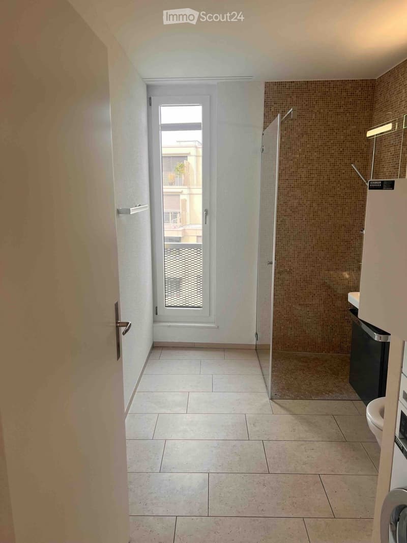 Modern bathroom with tiled floors and walls, featuring a toilet, sink, and a glass-enclosed shower stall. A window with a view to a balcony is visible.