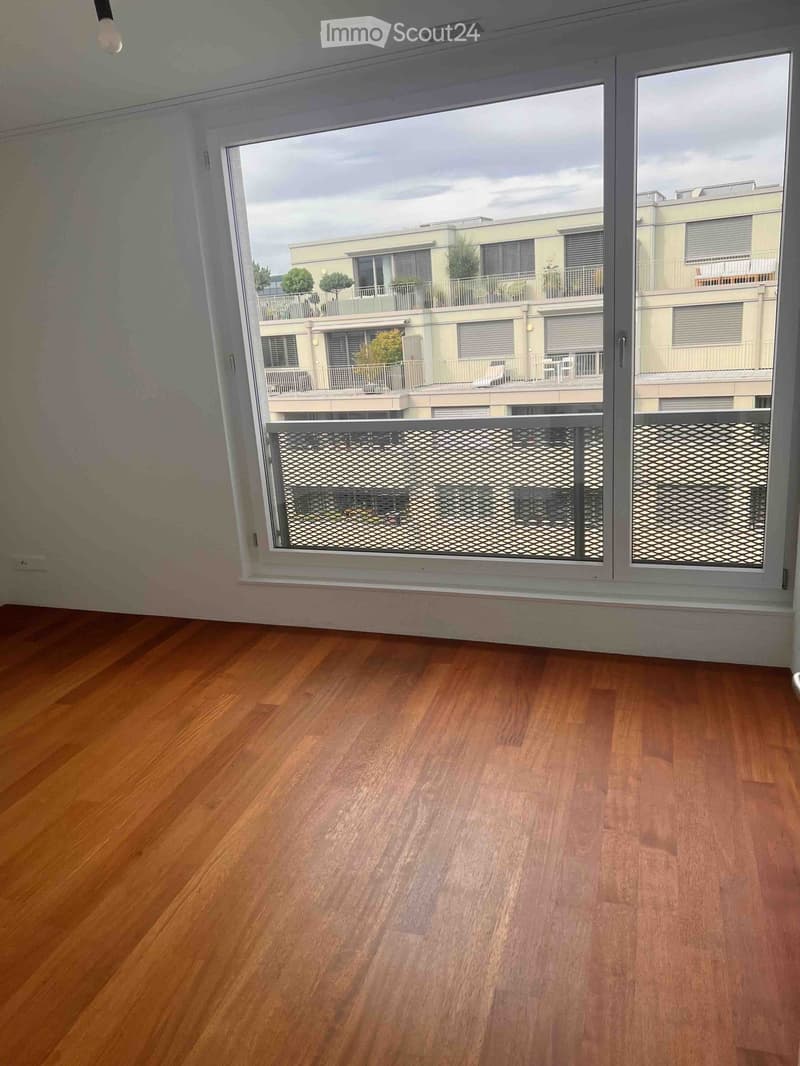 Empty room with wooden flooring and large windows offering a view of a multi-story building with balconies and greenery.